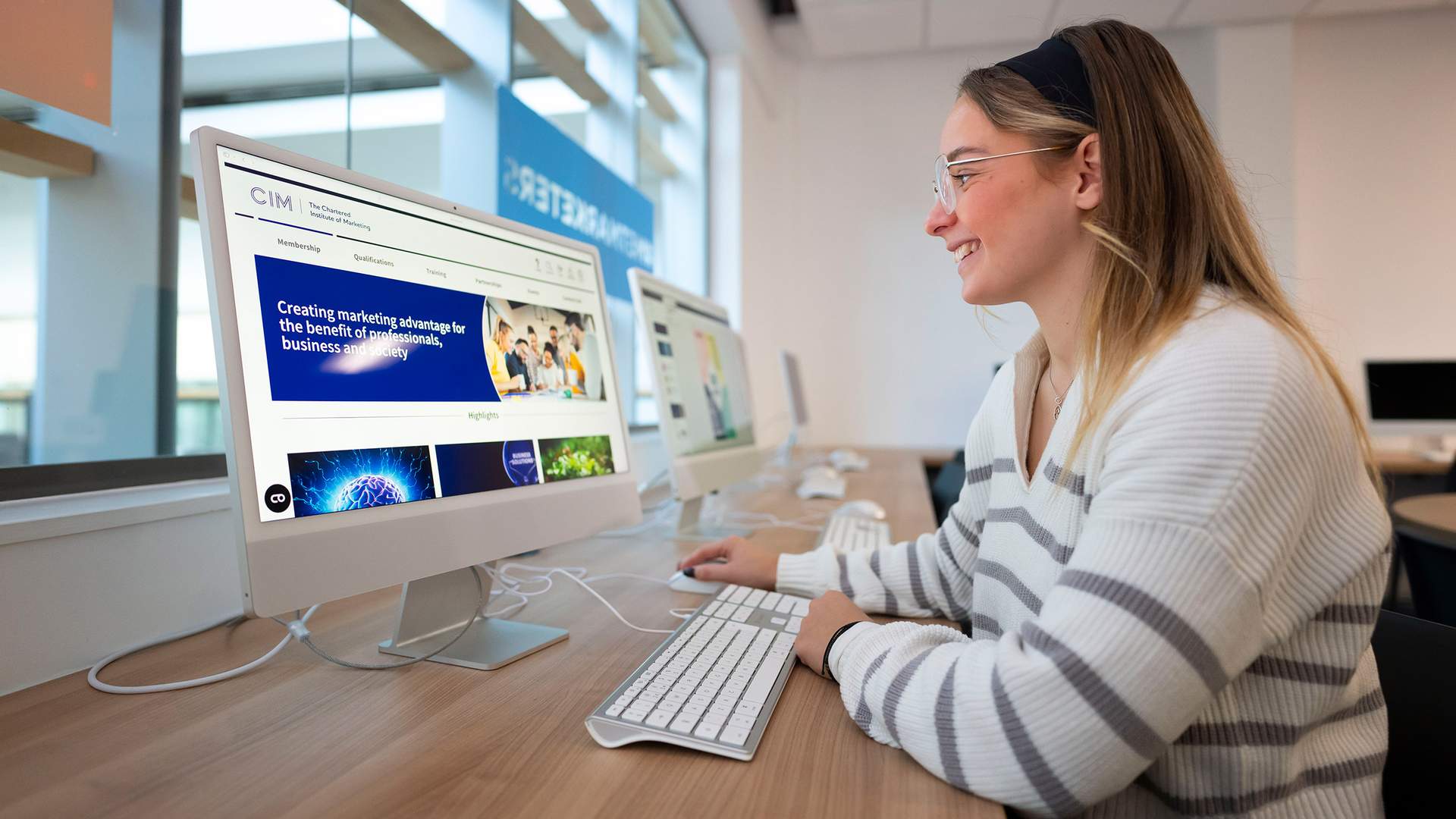 A person with long hair wearing a striped shirt is taking a selfie with a smartphone under a ring light. There are computers and potted plants in the background.