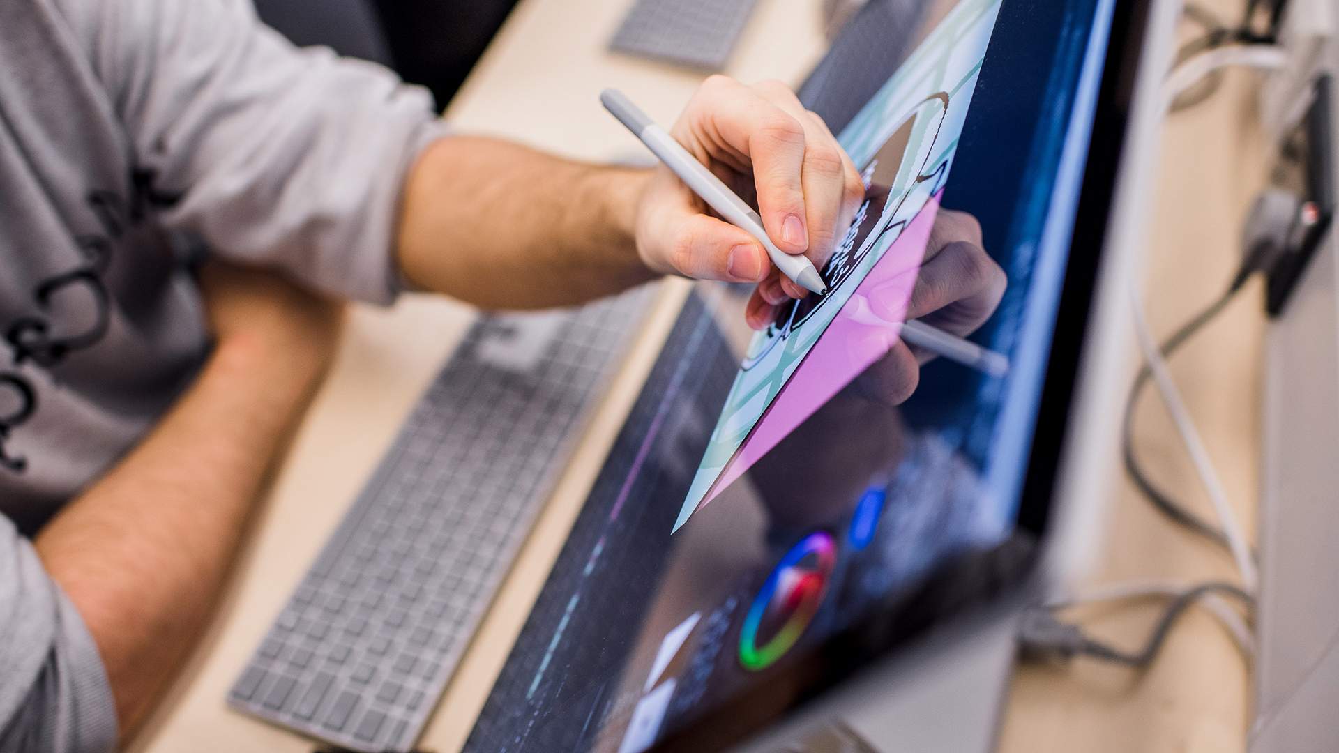 A close-up of a hand holding a stylus, drawing or writing on a digital tablet with a glowing screen. The background is blurred, creating a focused view on the tablet and the hand.