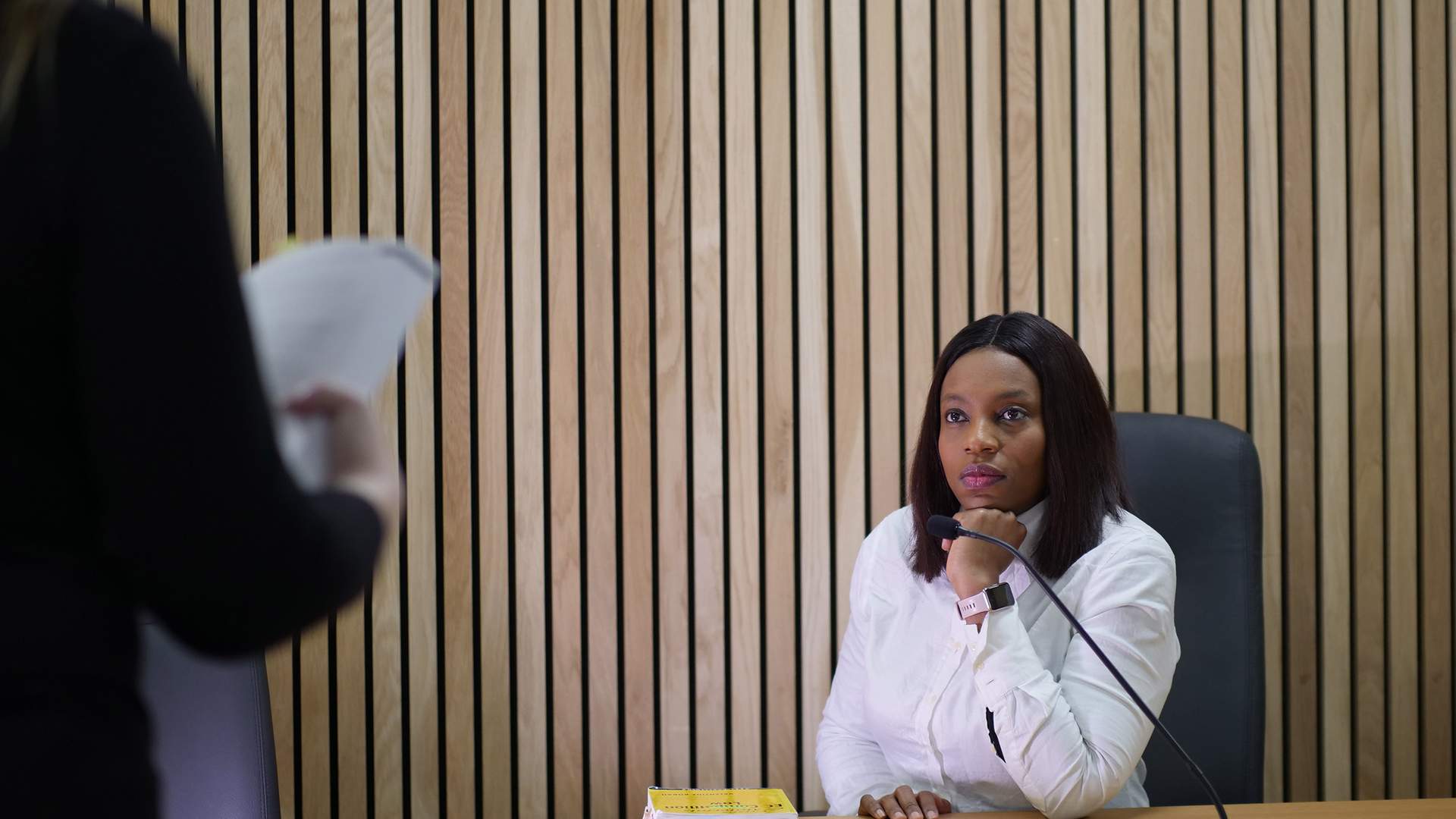 A woman seated at a wooden desk in a courtroom smiles and gestures with her hands while looking at a person sitting opposite her. The courtroom has wood-paneled walls and empty leather chairs.