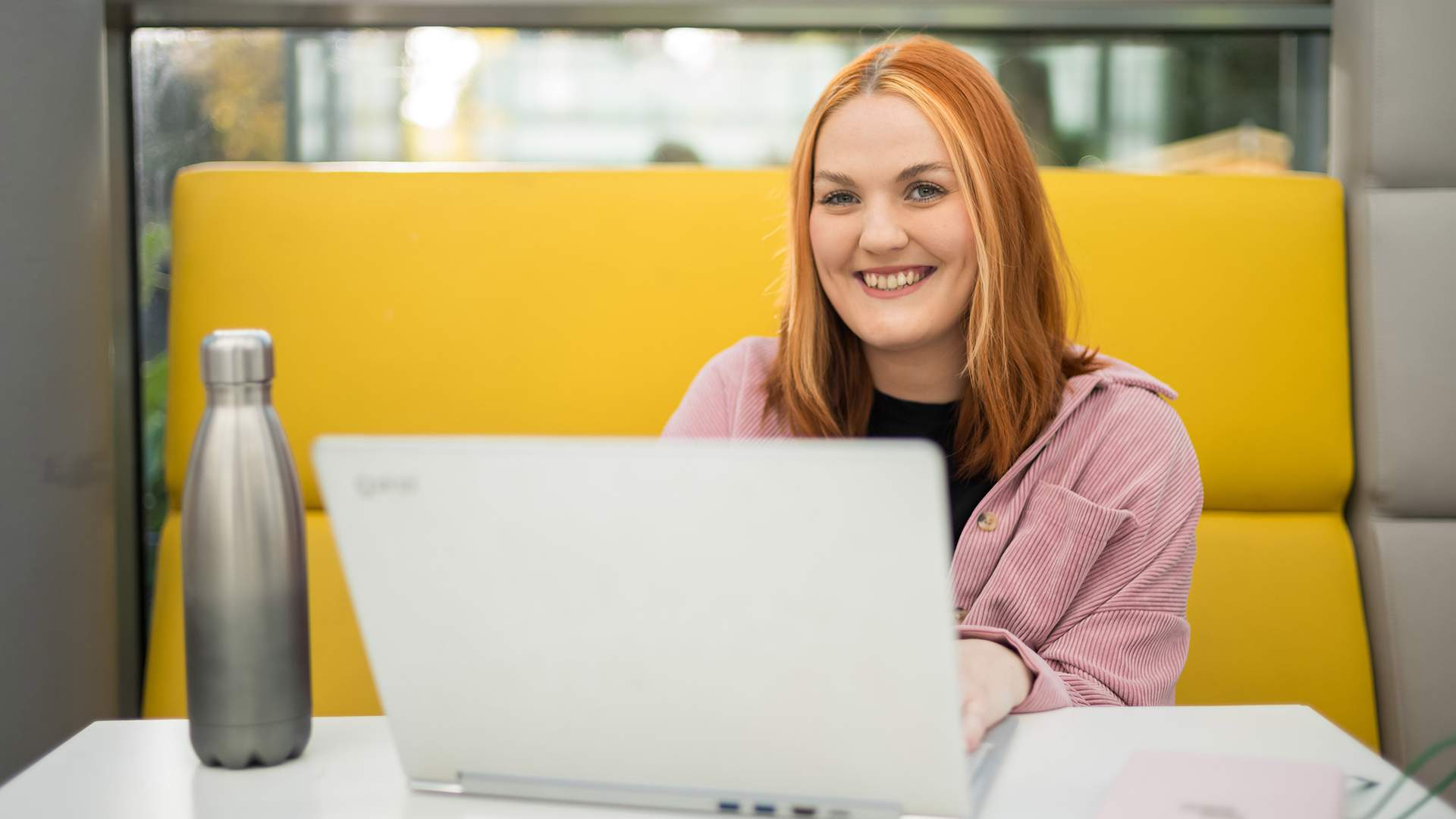 A woman with long brown hair and a striped shirt smiles while standing in an office. Theres a desk with a computer monitor and a plant in the background.