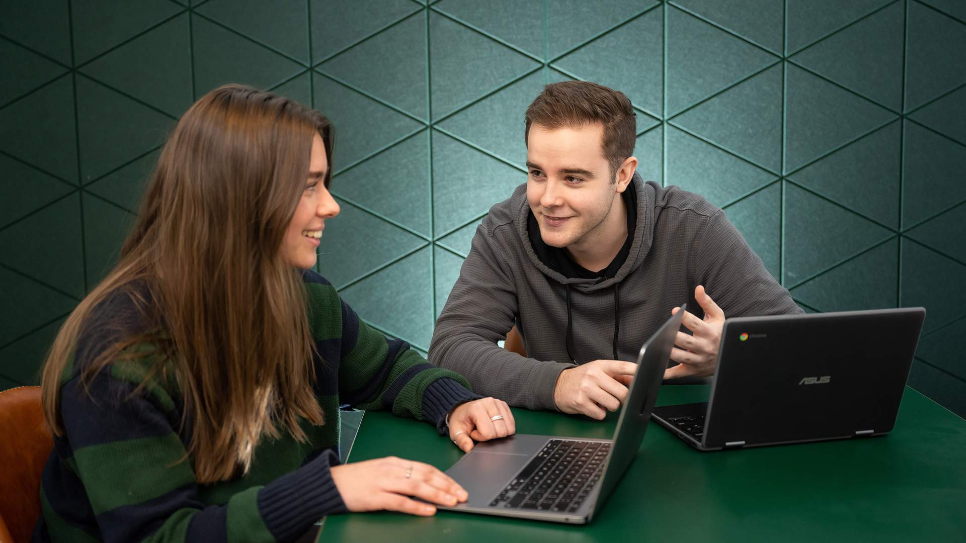 Two individuals seated at a table, each using a laptop, engaged in a collaborative work session.