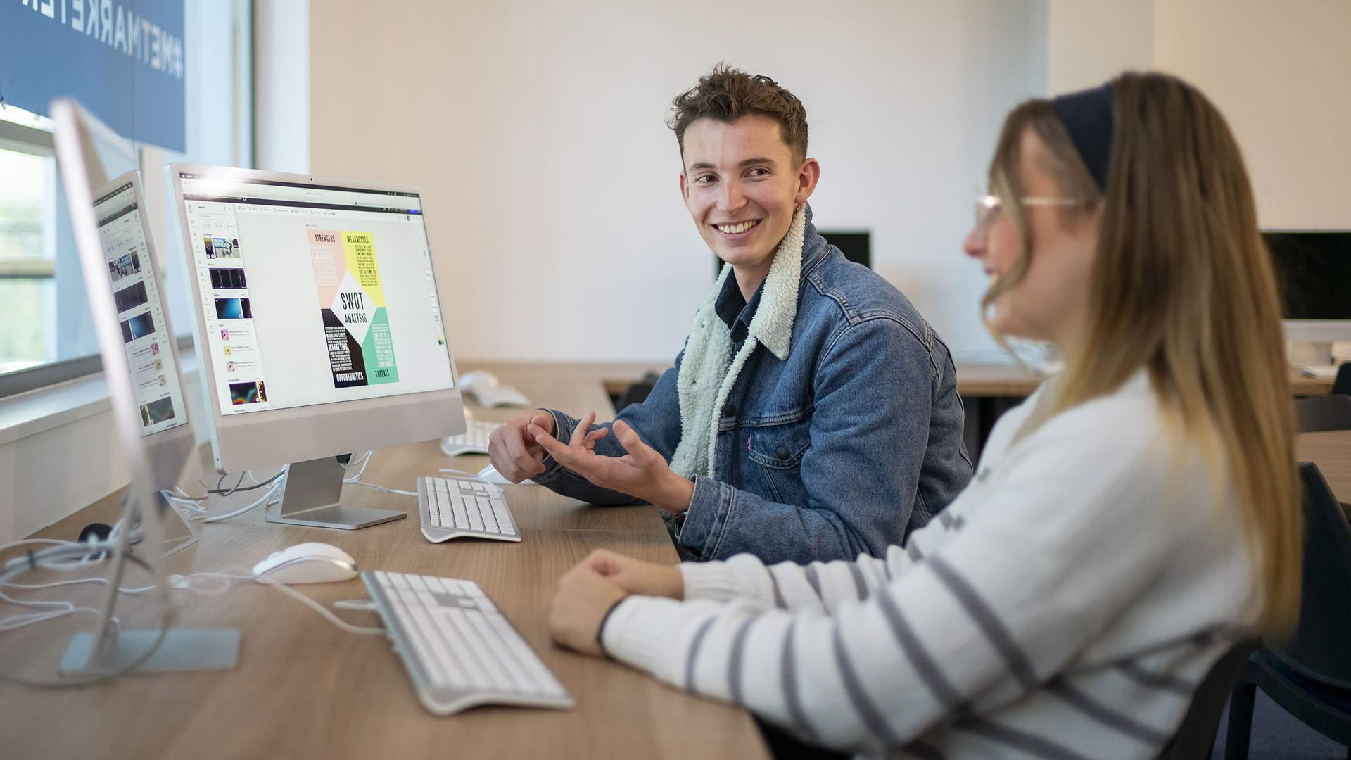 A person with long red hair is sitting at a table with a laptop and a water bottle. They are smiling and sitting in front of a large window with natural light. The background includes a yellow booth and indoor plants.