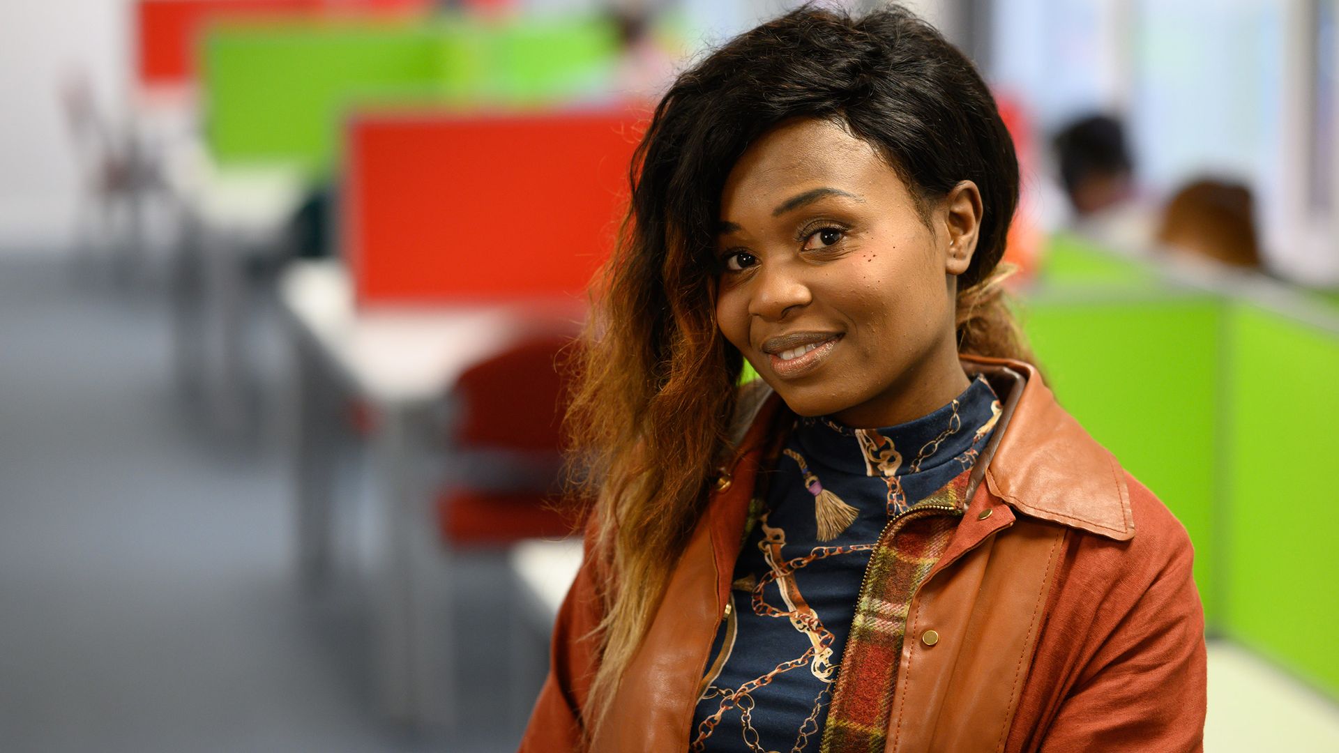 Two women are sitting and talking in an office setting. One woman with long hair and a patterned sweater smiles while the other, with hair tied back, faces her. Laptops are open on the table in front of them.