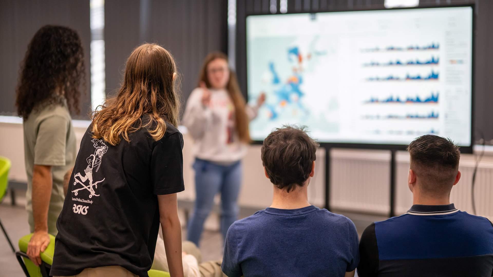 Two people are focused on a computer screen, working together. The woman on the left is wearing a floral blouse, and the man on the right is in a black jacket. They are in a bright room with other computer equipment visible in the background.