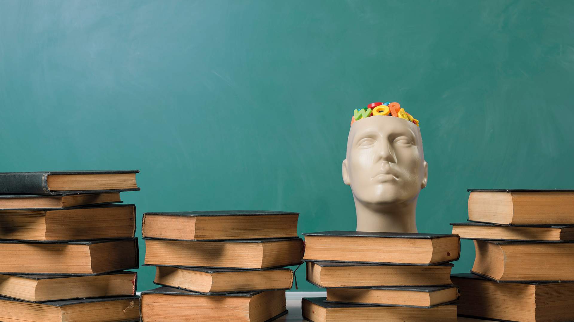 A person with curly hair is sitting at a table, smiling and writing in a notebook with a pen. They are wearing a gray sweater. The background features vertical wooden slats and a window.