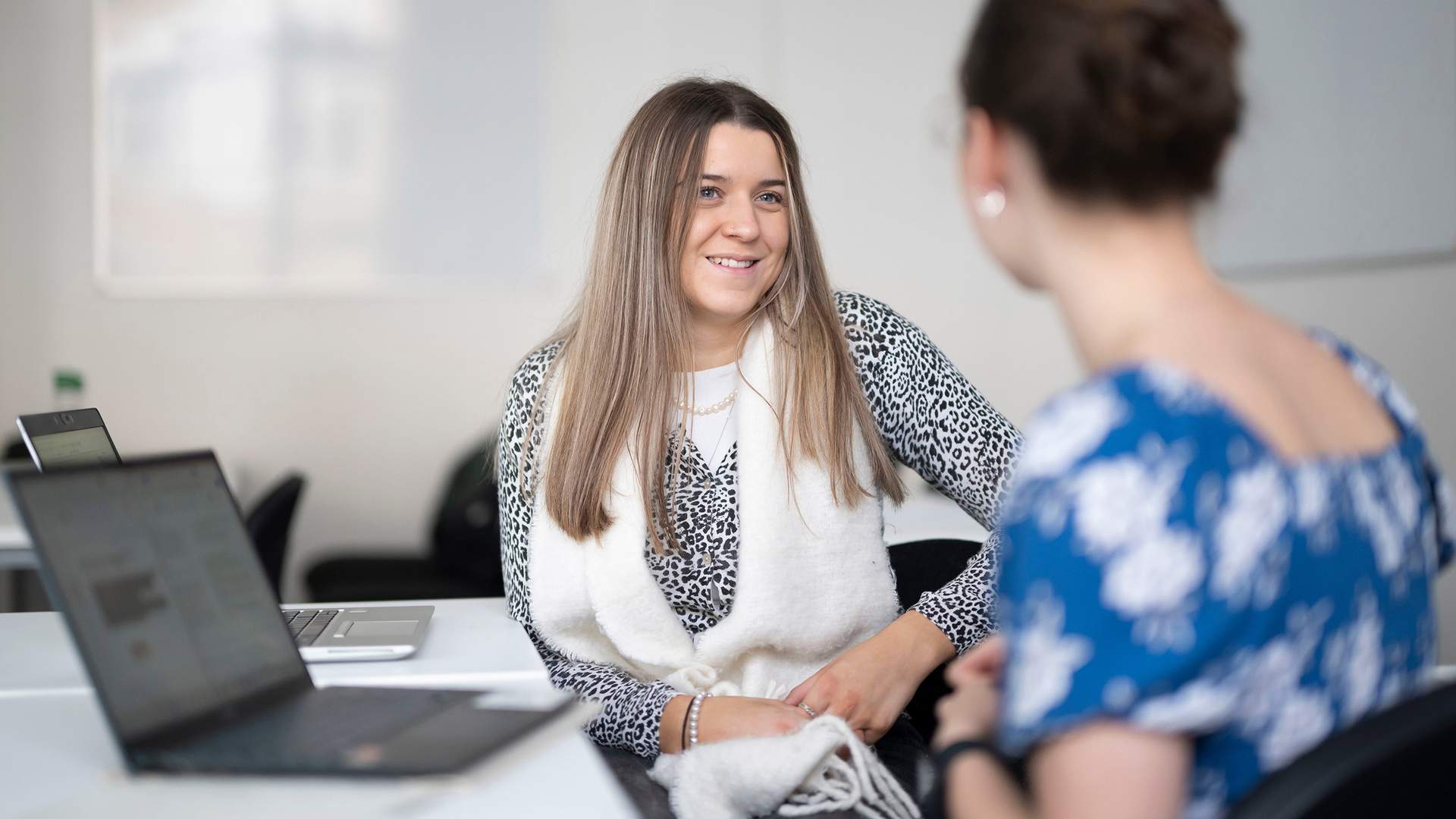 Two women sitting on a couch, one holding a clipboard and pen, smiling warmly at each other. The setting is a bright, cozy living room with a plant and artwork in the background.