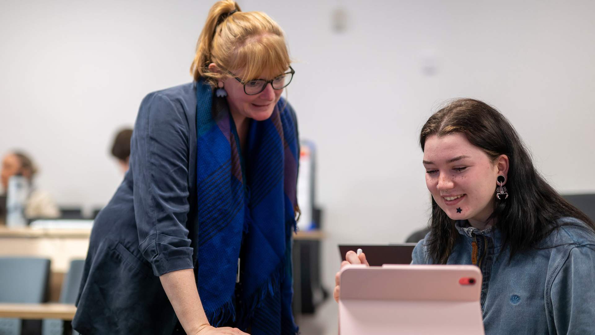 Two people are seated and talking at a table. One person in a green top and headscarf is smiling while using a laptop. The other, with curly hair, is seen from behind. They appear to be engaged in a positive conversation in an office setting.