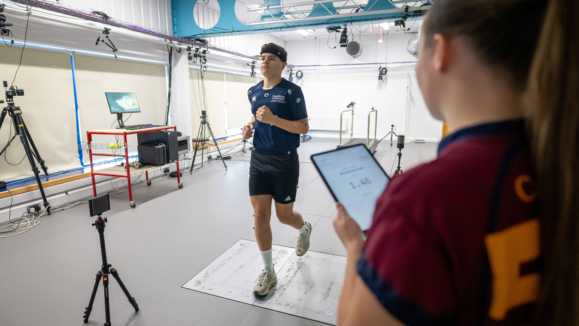 A person runs on a treadmill in a technology-equipped lab, surrounded by cameras and a computer screen. A bright green light is visible in the foreground. The setting suggests a motion analysis or biomechanics study.