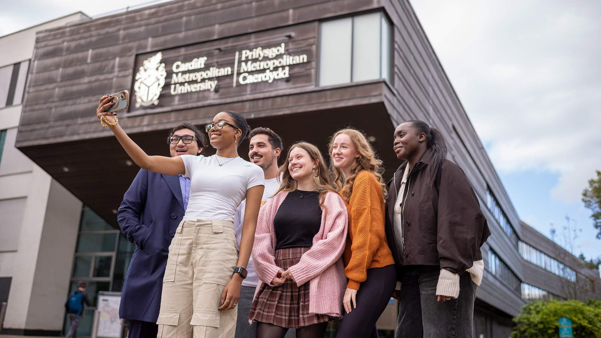 A group of four people walking and talking outdoors, holding coffee cups. Theyre dressed in casual winter clothing, with trees and modern buildings in the background under a partly cloudy sky.