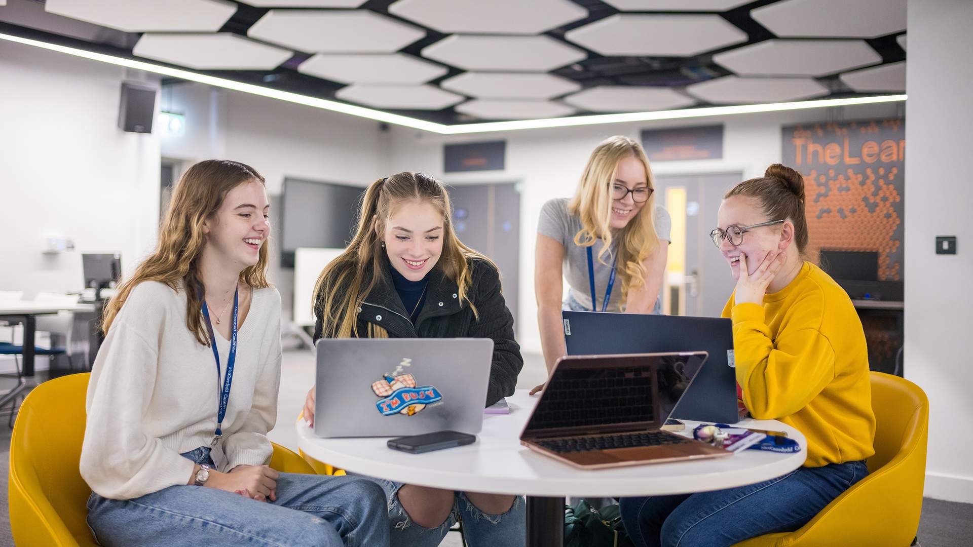 A person wearing glasses and a black t-shirt is sitting on a bright green chair, laughing. Another person with shoulder-length hair is in the foreground, slightly out of focus. The setting appears to be a casual indoor environment.