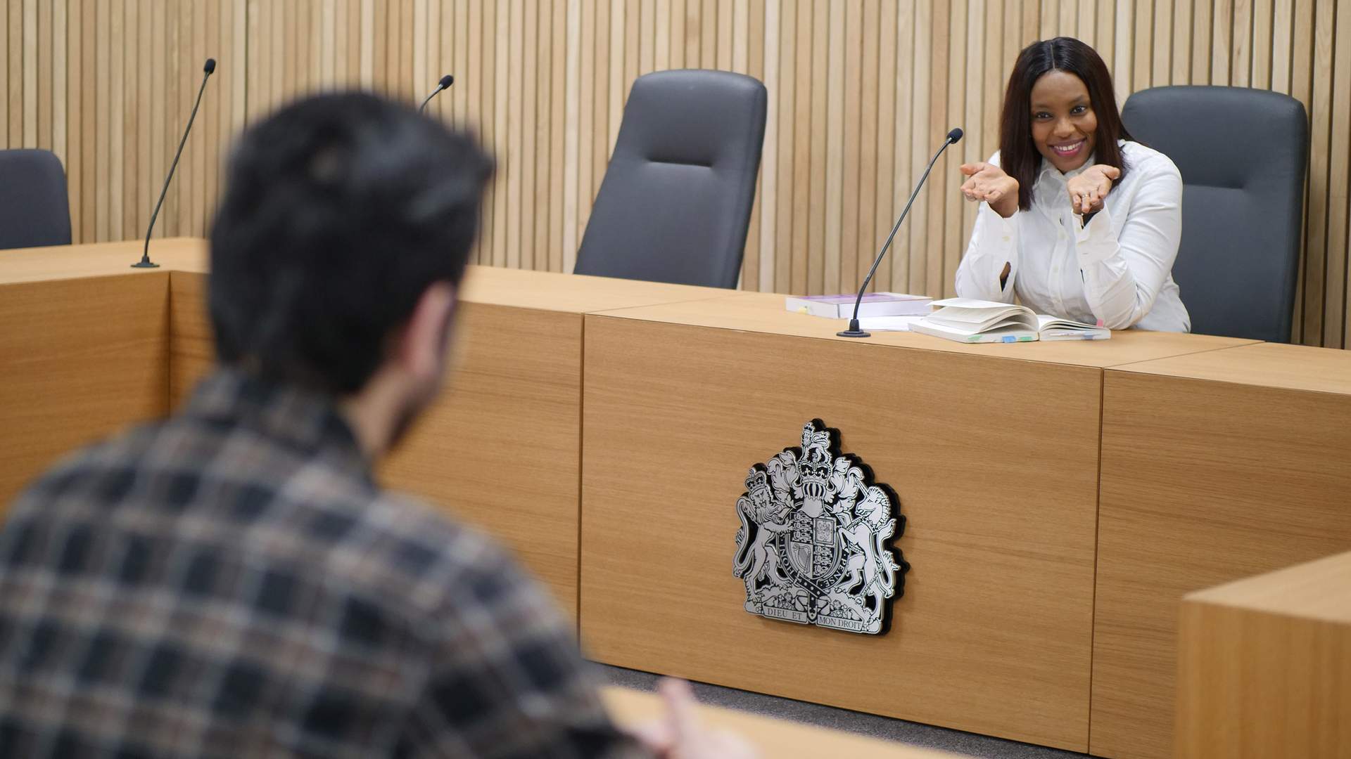 A man in a light gray suit and white shirt is sitting at a table, smiling and gesturing with his hands. He faces a person with long hair, seated across from him. A glass of water and papers are on the wooden table.