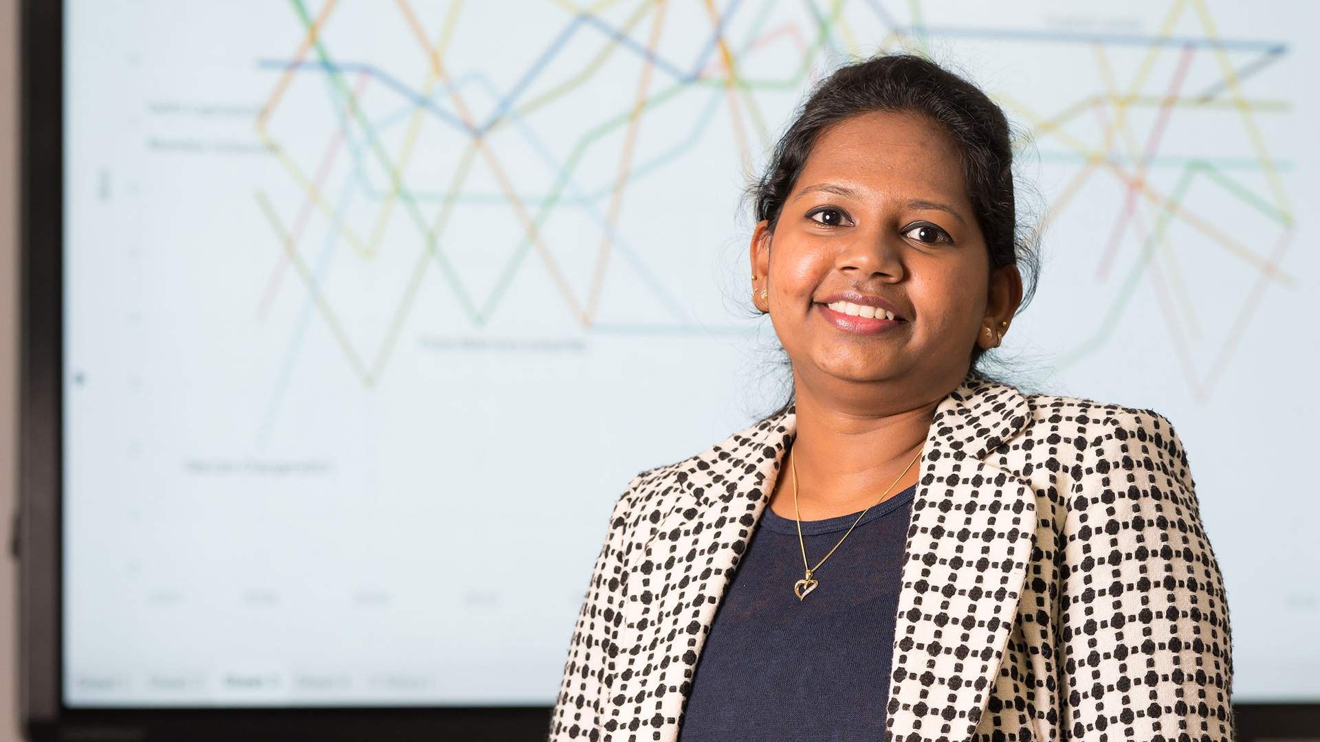 A woman with long dark hair sits on a bright green couch, smiling while using a laptop covered in various black and white stickers. She wears a black dress, and the background features a modern, minimalist office setting.