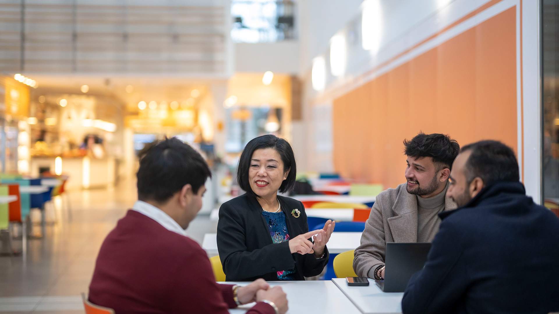 A person in a black blazer stands smiling in front of a modern building with Cardiff Metropolitan University signage. The sky is clear.