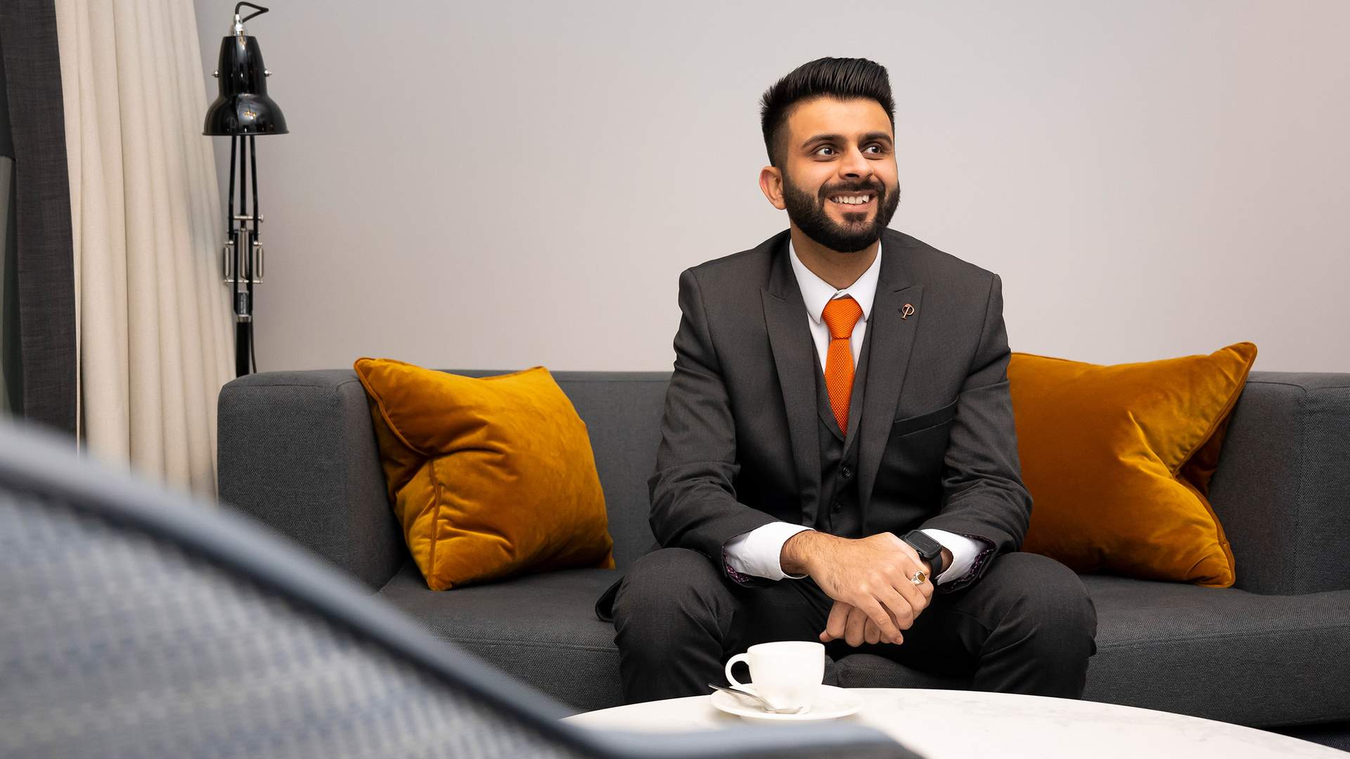 A man wearing a dark suit with an orange tie stands smiling in a modern office lobby with wooden floors and open double glass doors in the background. An orchid is visible on a table to the left.