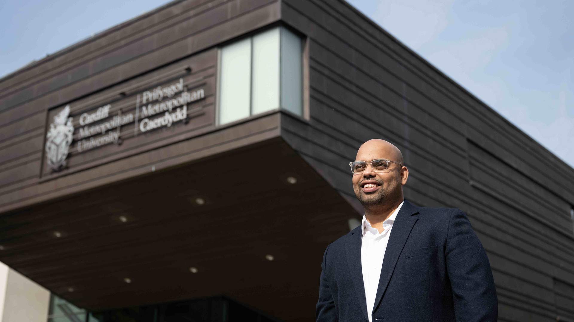 A person with glasses and a mustache, wearing a green jacket, is sitting outdoors at a table with a laptop. Theres a blue mug and a pen next to the laptop. Trees and foliage are visible in the blurred background.