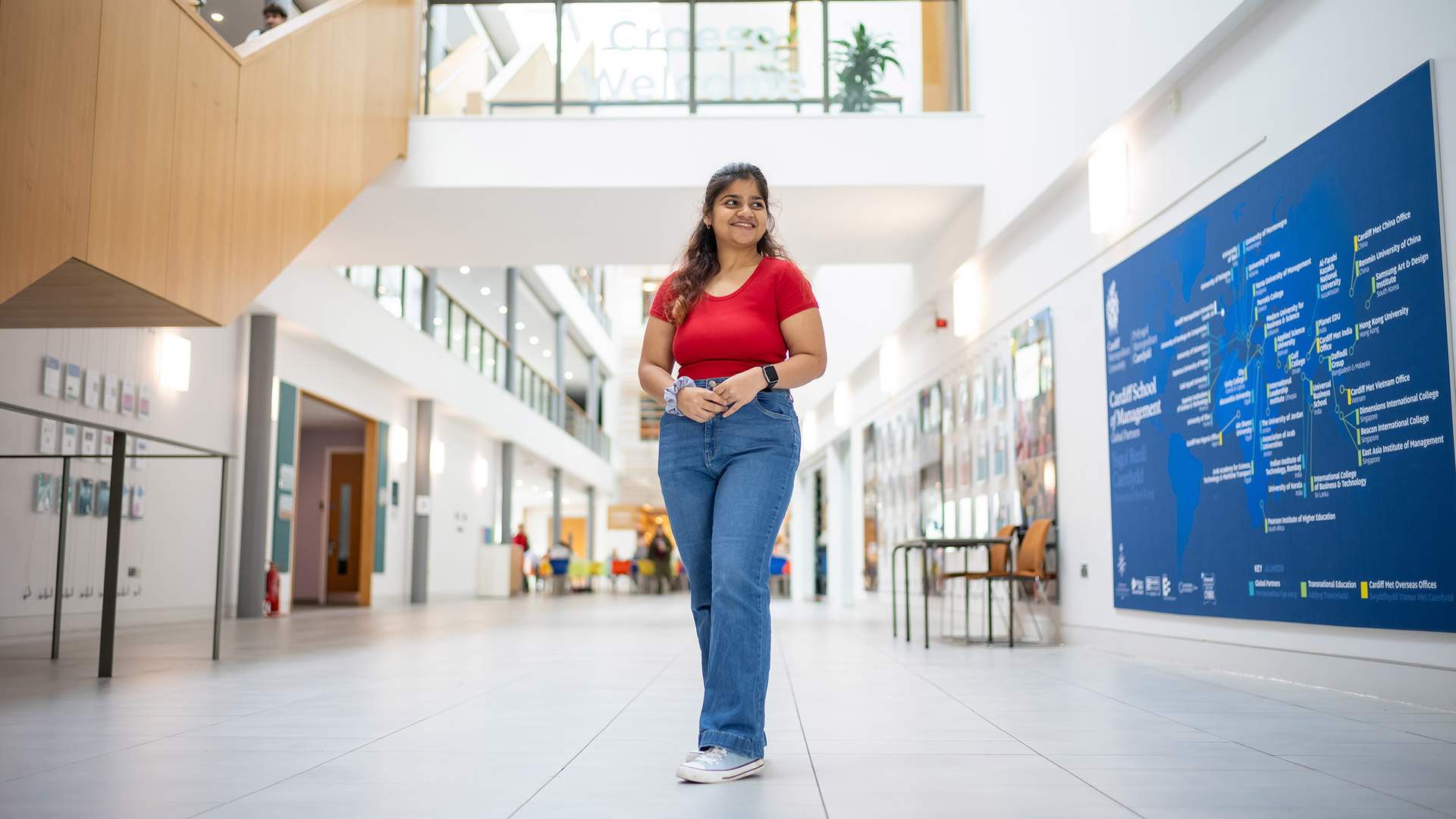 A man in a white shirt stands with his hands on a railing, smiling broadly in a modern, multi-story building with glass windows and wooden accents.