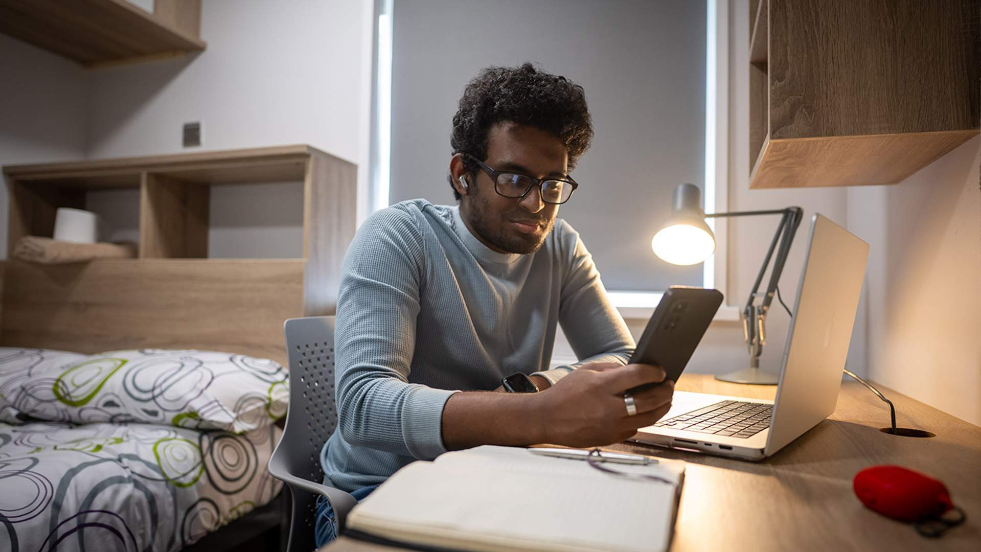 A man seated at a desk, working on a laptop while holding a cell phone in his hand.