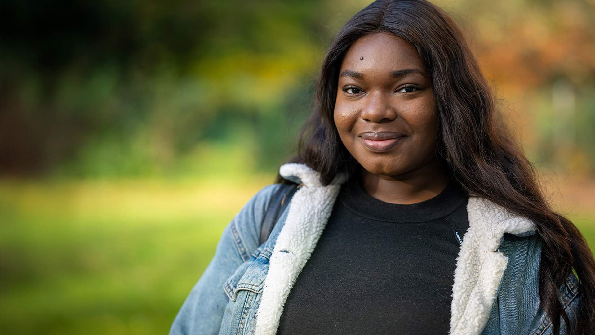 A young woman wearing a black shirt and a denim jacket, smiling confidently against a neutral background.