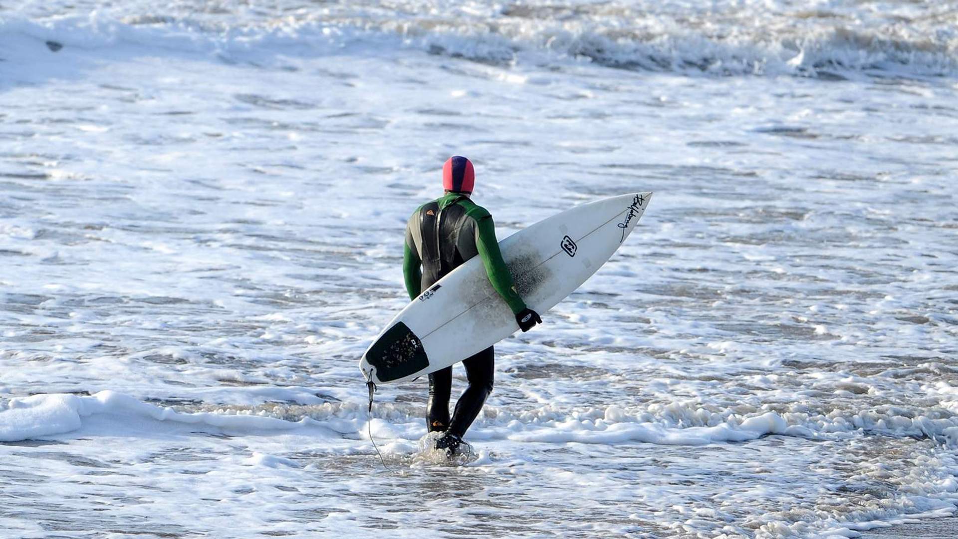 A surfer wearing a a wetsuit and carrying a surfboard walks through the waves on a beach.