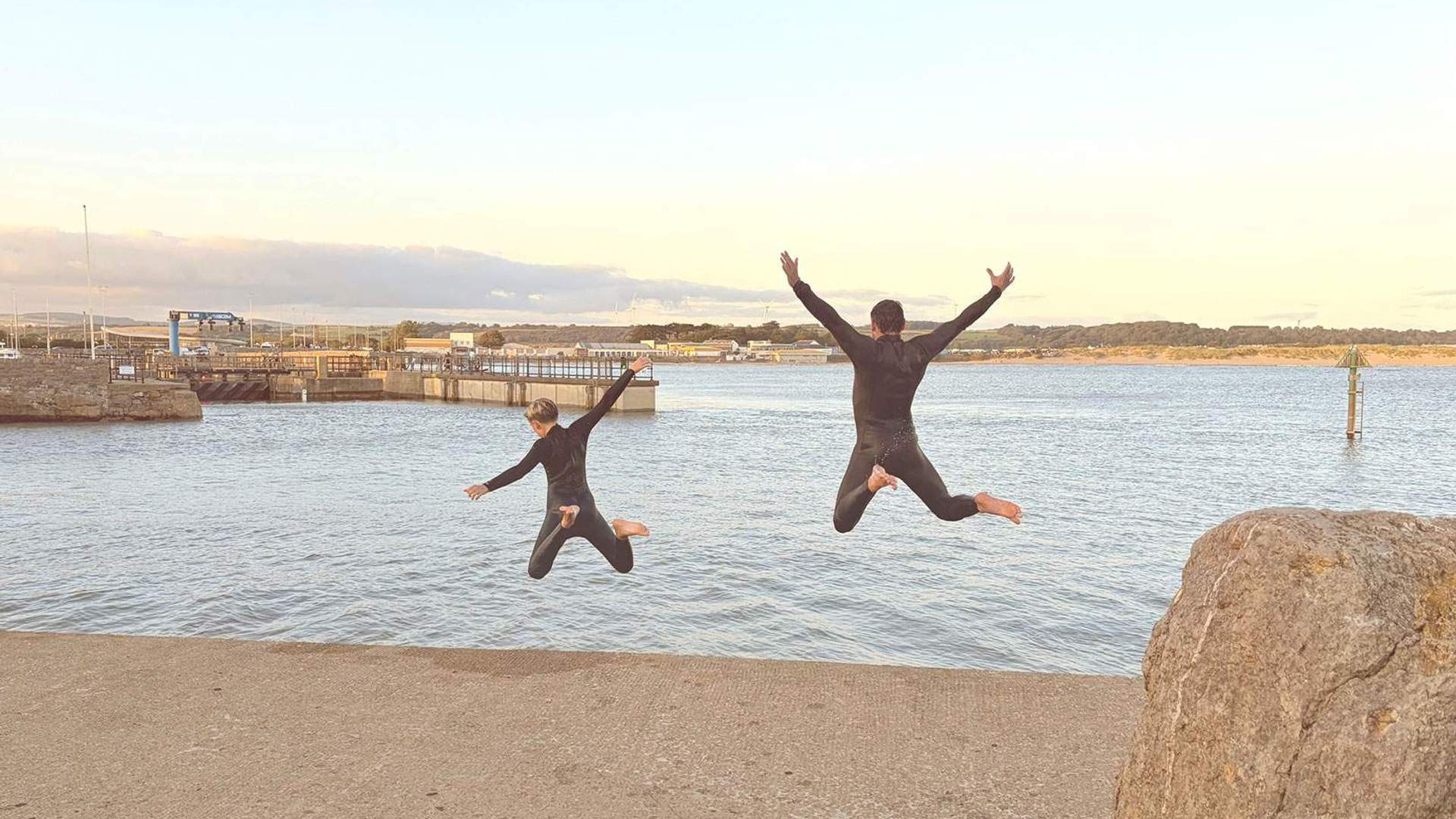 Two people wearing wetsuits jump into the water from a concrete dock