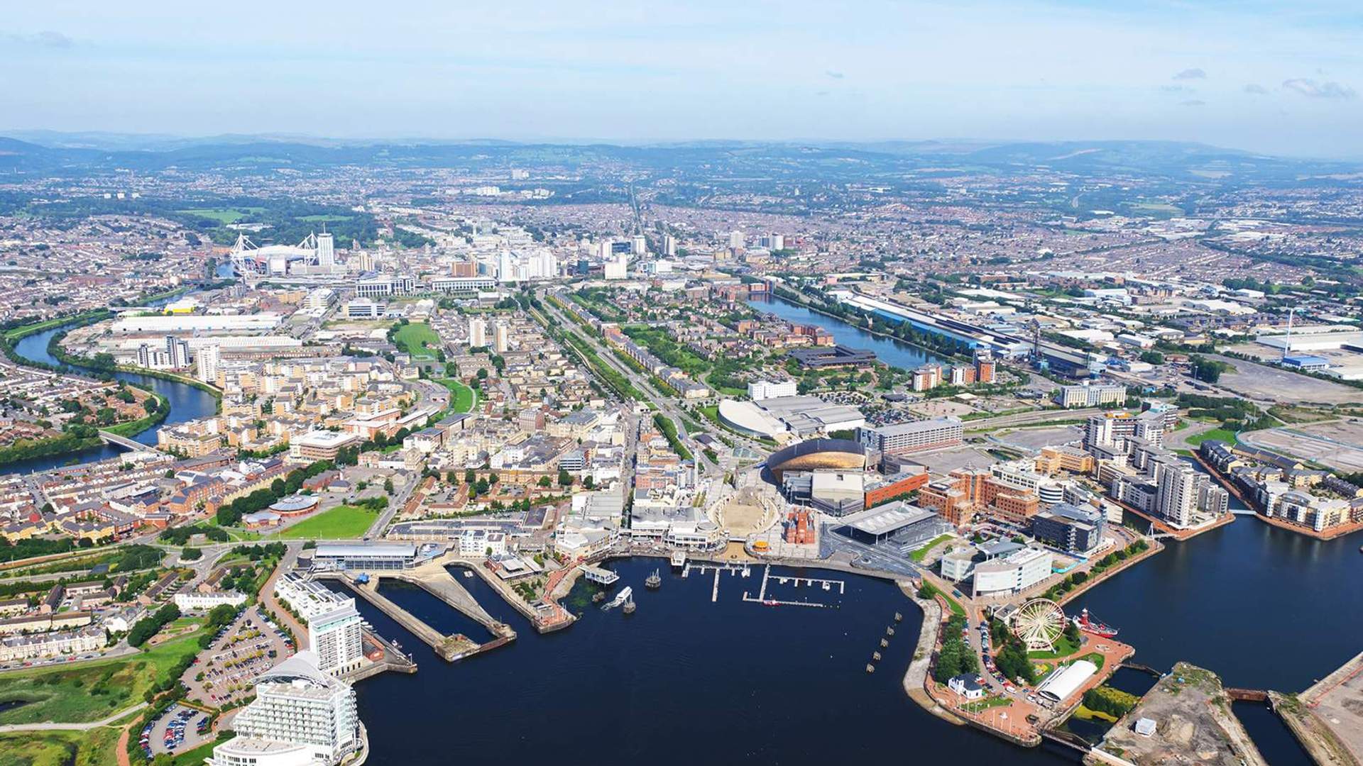 Aerial view of a vibrant waterfront city with winding rivers, numerous buildings, and a marina. The landscape features green spaces and urban areas under a clear blue sky.