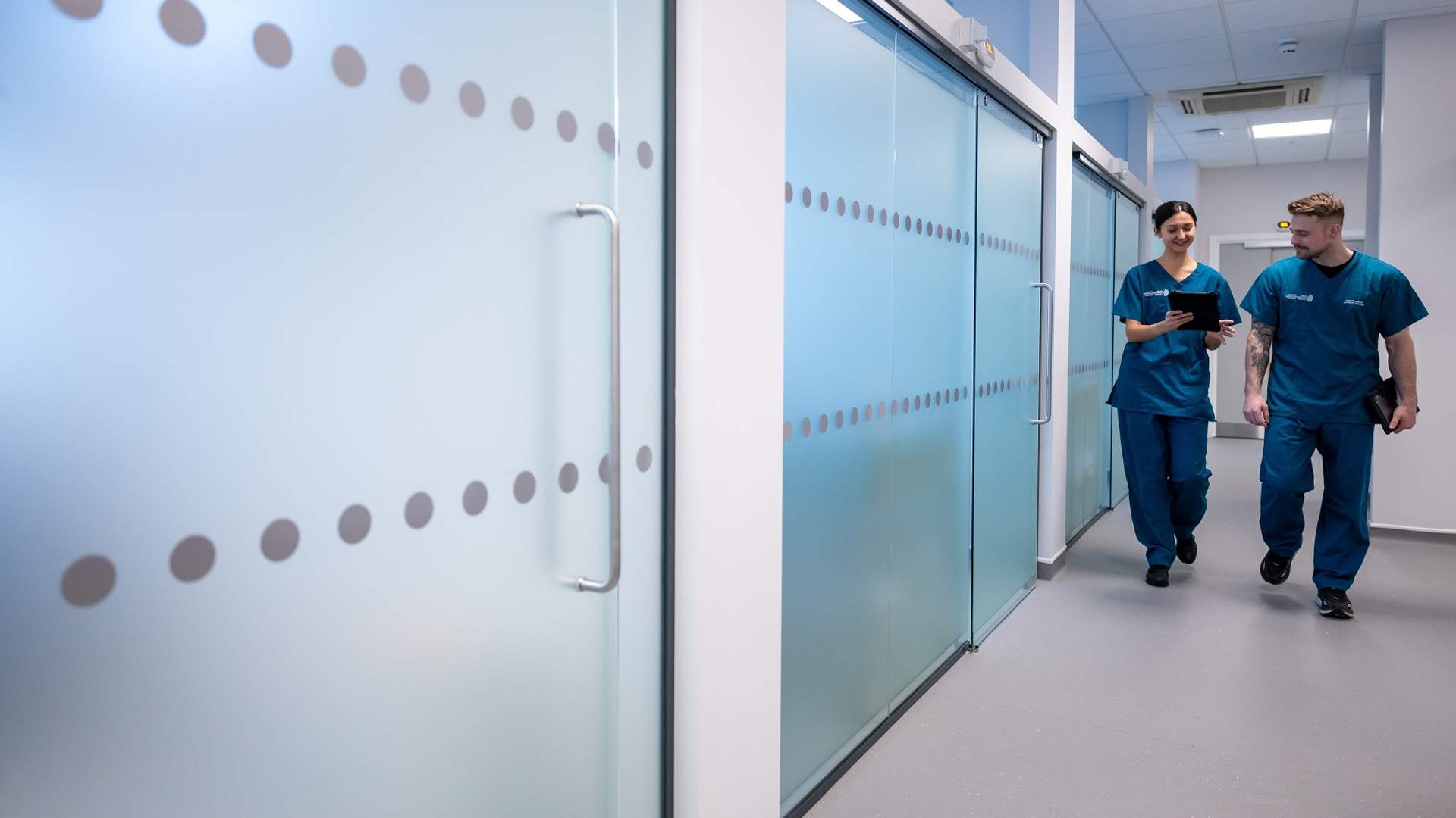 Two young adults in blue nursing scrubs walk through a corridor