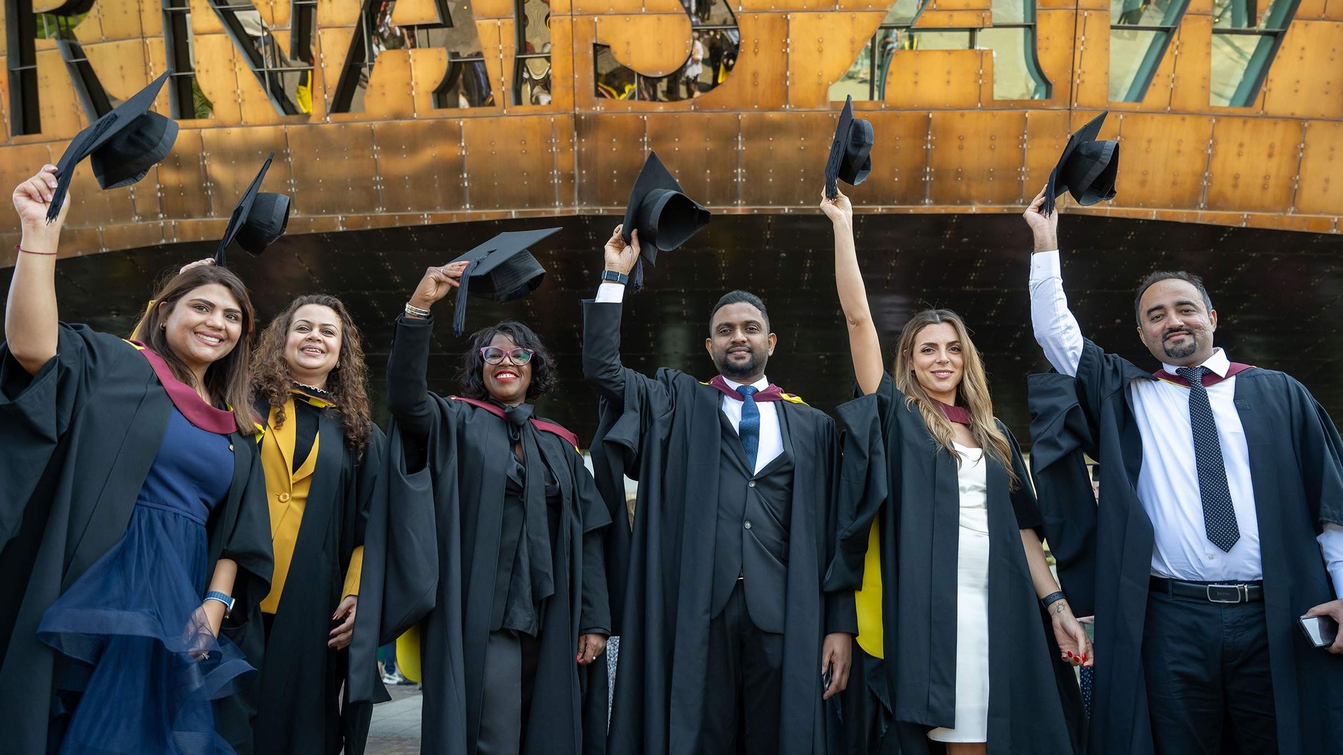 Six students in graduation gowns line up in front of the Wales Millennium Centre, holding their caps in the air.