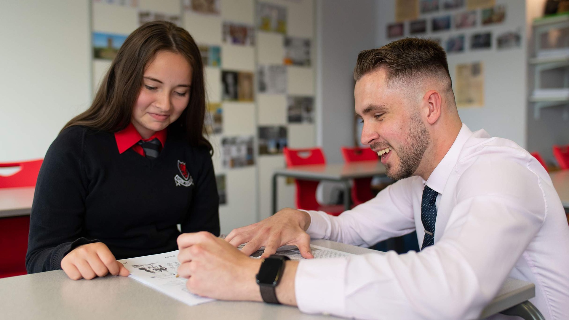 A teacher kneels next to a pupil sitting at a desk, pointing to the open page of a book.