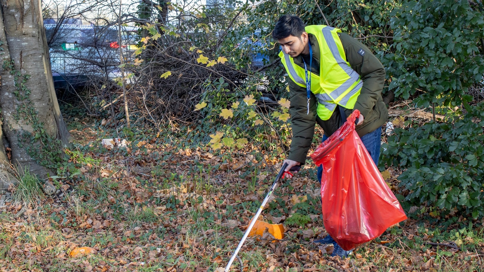 Young adult in high visibility jacket uses a litter picker to collect rubbish in a large red bin bag