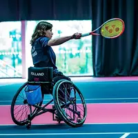 A female wheelchair tennis player on an indoor court with her arm outstretched holding a tennis racket.