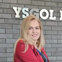 A woman with blonde hair wearing a red jacket. Behind her is a brick wall with the lettering YSGOL on it.