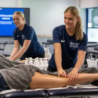 Two women in blue shirts providing leg massages in a class room setting.