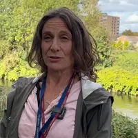 A woman in a pink shirt stands beside a river, surrounded by greenery and reflecting sunlight on the water's surface.