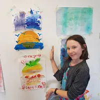 A young girl stands in front of a wall displaying various colorful paintings.