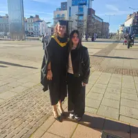 A women and her young daughter after graduation and in her gown stands on a brick walkway, celebrating their achievement with smiles and pride.