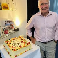 A man stands beside a decorated cake, smiling and celebrating receiving his OBE.