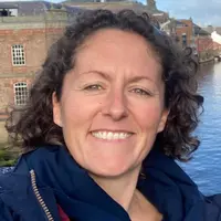 A woman with curly hair in a blue jacket stands by a river, with water flowing gently in the background.