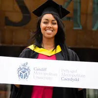 A Cardiff Met student, in graduation cap and gown, stands holding a sign featuring the Cardiff Met logo and name