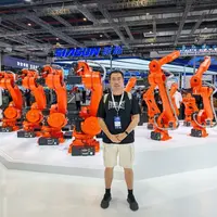 At an exhibition, a man poses in front of several orange robots, highlighting advancements in robotics and automation.