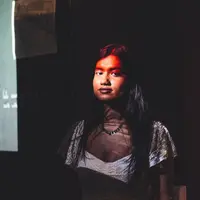 A young woman looks at the camera in a low light room, illuminated by the light from a projector
