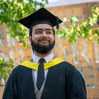 A man wearing a graduation cap and gown. In the background is Wales Millennium Centre.