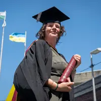 A woman wearing a graduation cap and gown pictured against a bright blue sky. In the background are flag poles with the flags of Wales and Ukraine.
