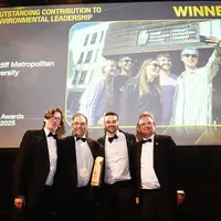 Cardiff Met staff accept an environmental leadership award on stage beneath a winner announcement screen.