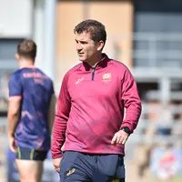 A man wearing a red shirt and blue shorts strolls on a green field, coaching rugby players.