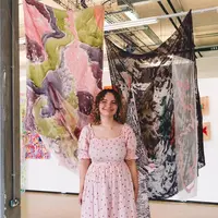 A young woman in a light pink dress stands in front of two large patterned silks