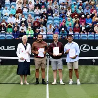 Four people stand in front of a tennis net on a court in front of a large crowd of spectators