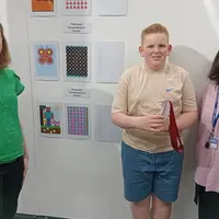 A young boy holding an award stands in front of a display of computer generated artwork. Standing on either side of him are staff from Cardiff Met.
