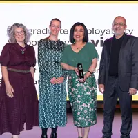 A group of four people pose for a photo in front of large display screen. One of them is holding an award in their hands.