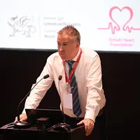 Professor Barry McDonnell standing at a lectern delivering a presentation. On the screen behind him are the logos of Welsh Government and British Heart Foundation.