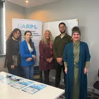 A diverse group of people stand next to a table. A banner with DARPL on it is visible in the background.